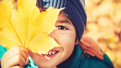Young boy in autumn, wearing woolly hat with his face partially obscured by a leaf that he is holding.
