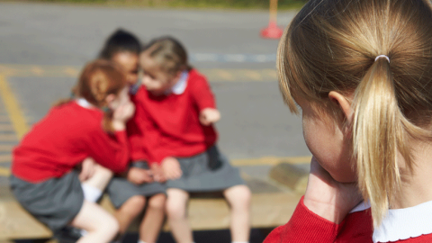 Close-up of the back of the head of a blonde primary school girl watching three little girls talking and poking fingers in her direction