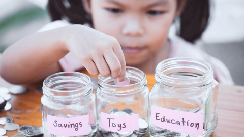 Close-up of little Asian girl dispensing coins into different glass saving jars labelled Savings, Toys and Education.