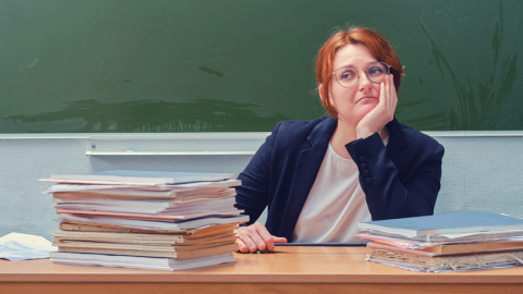 Despondent-looking red-haired teacher sitting at desk piled high with books in front of a blackboard.
