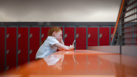 Red-haired schoolgirl sitting alone looking at her phone in front of school lockers