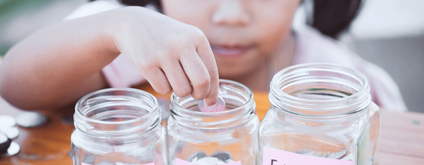 Close-up of little Asian girl dispensing coins into different glass saving jars labelled Savings, Toys and Education.