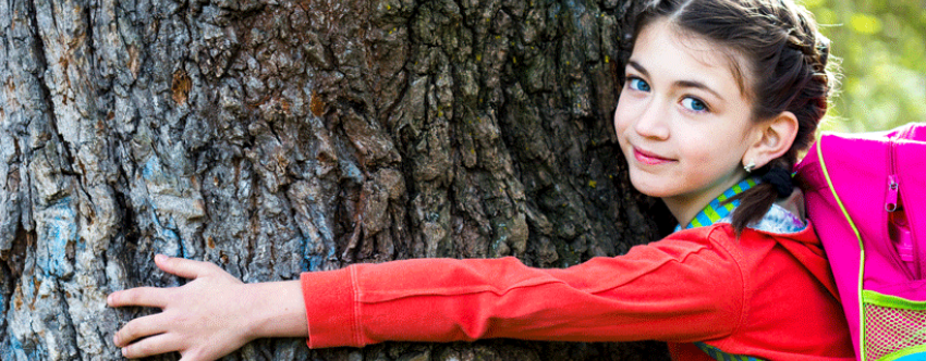 Little girl in a red coat wearing a pink backpack, looking happy hugging a tree