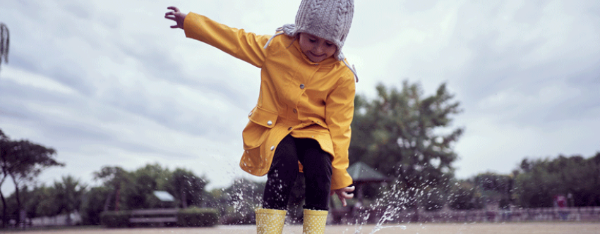 Little girl dressed in a yellow waterproof coat, hat and wellies, joyously jumping in a puddle. 