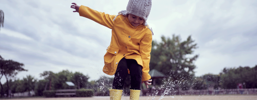 Little girl dressed in a yellow waterproof coat, hat and wellies, joyously jumping in a puddle.