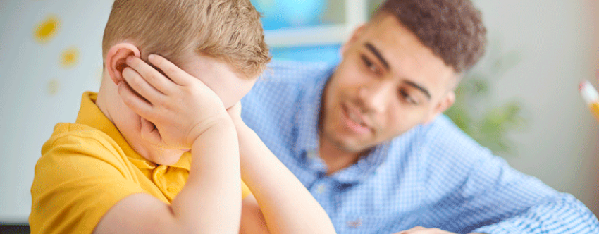 Male teacher comforting small blond school boy who looks distressed.