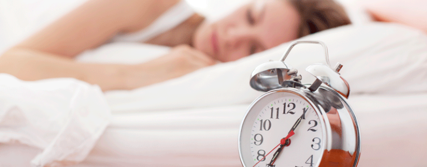 Alarm clock in the foreground with a woman asleep behind it.