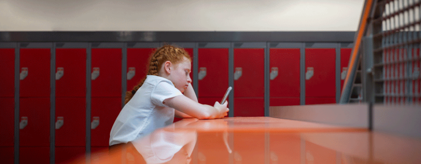 Red-haired schoolgirl sitting alone looking at her phone in front of school lockers